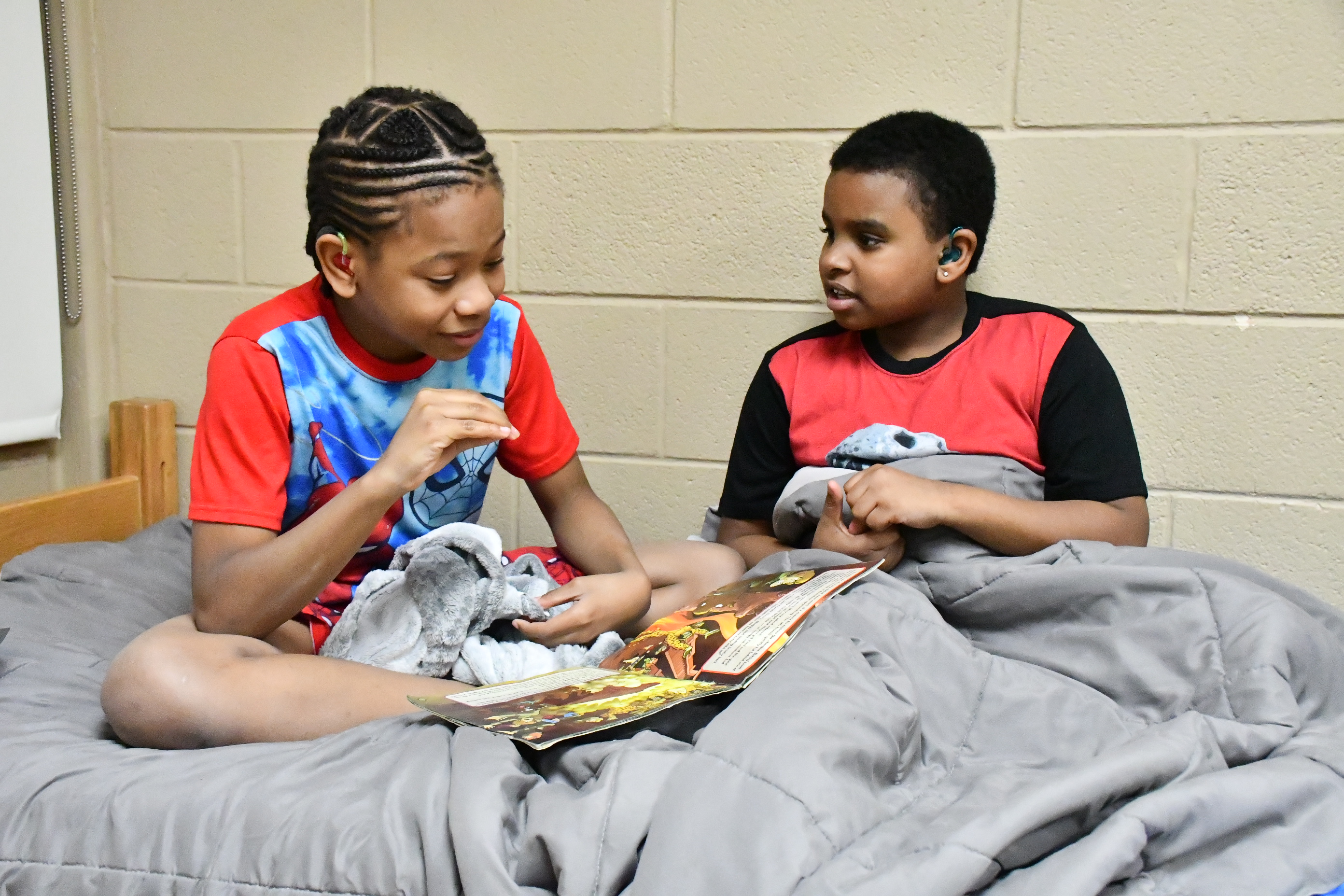 Two boys sit in a bed with a book in their lap while one signs to the other in ASL.