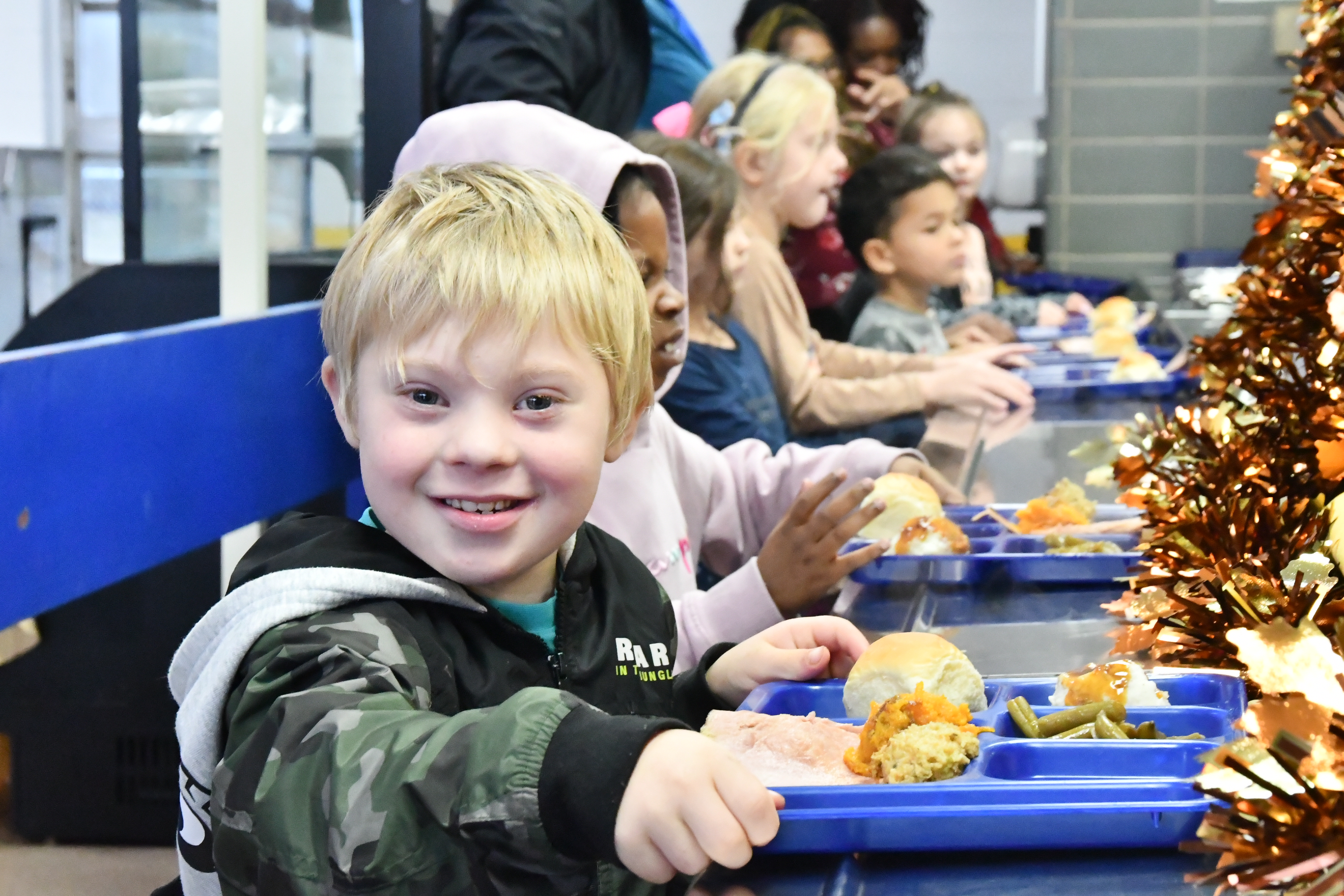 A young boy stands in a lunch line smiling while holding a tray of food.