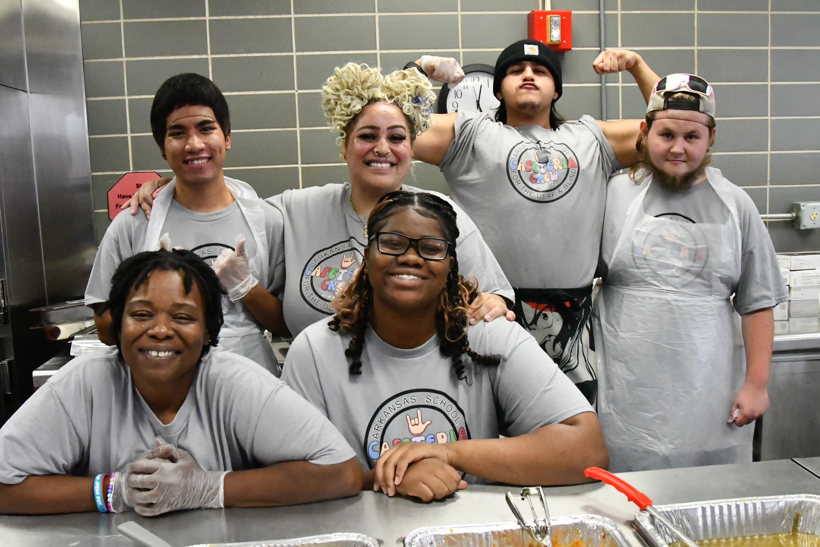 The cafeteria crew of six stands behind the food line smiling and posing.