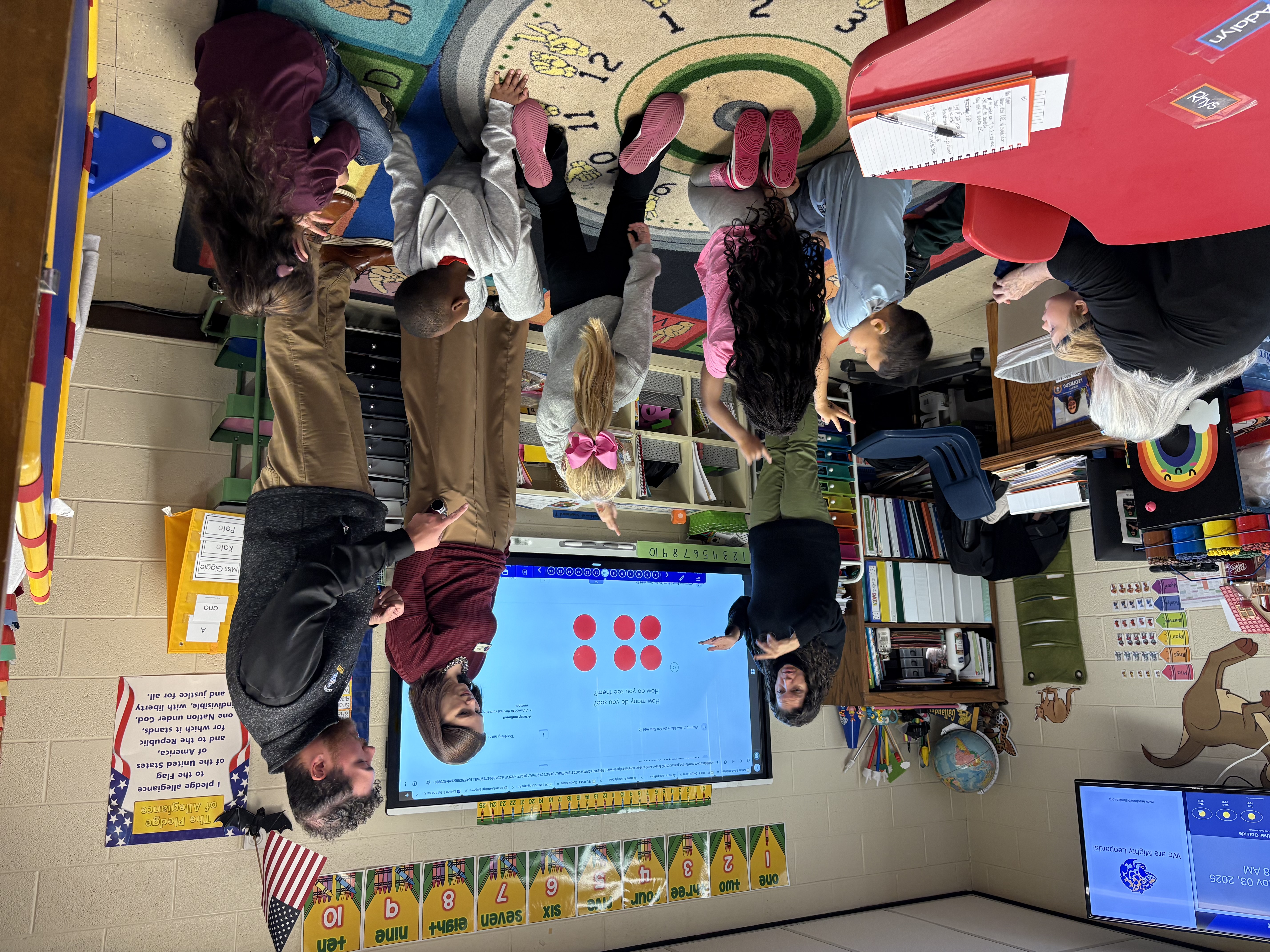 Three adults stand in front of a Smart Board instructing while several children on the floor have their hands up in varying gestures.