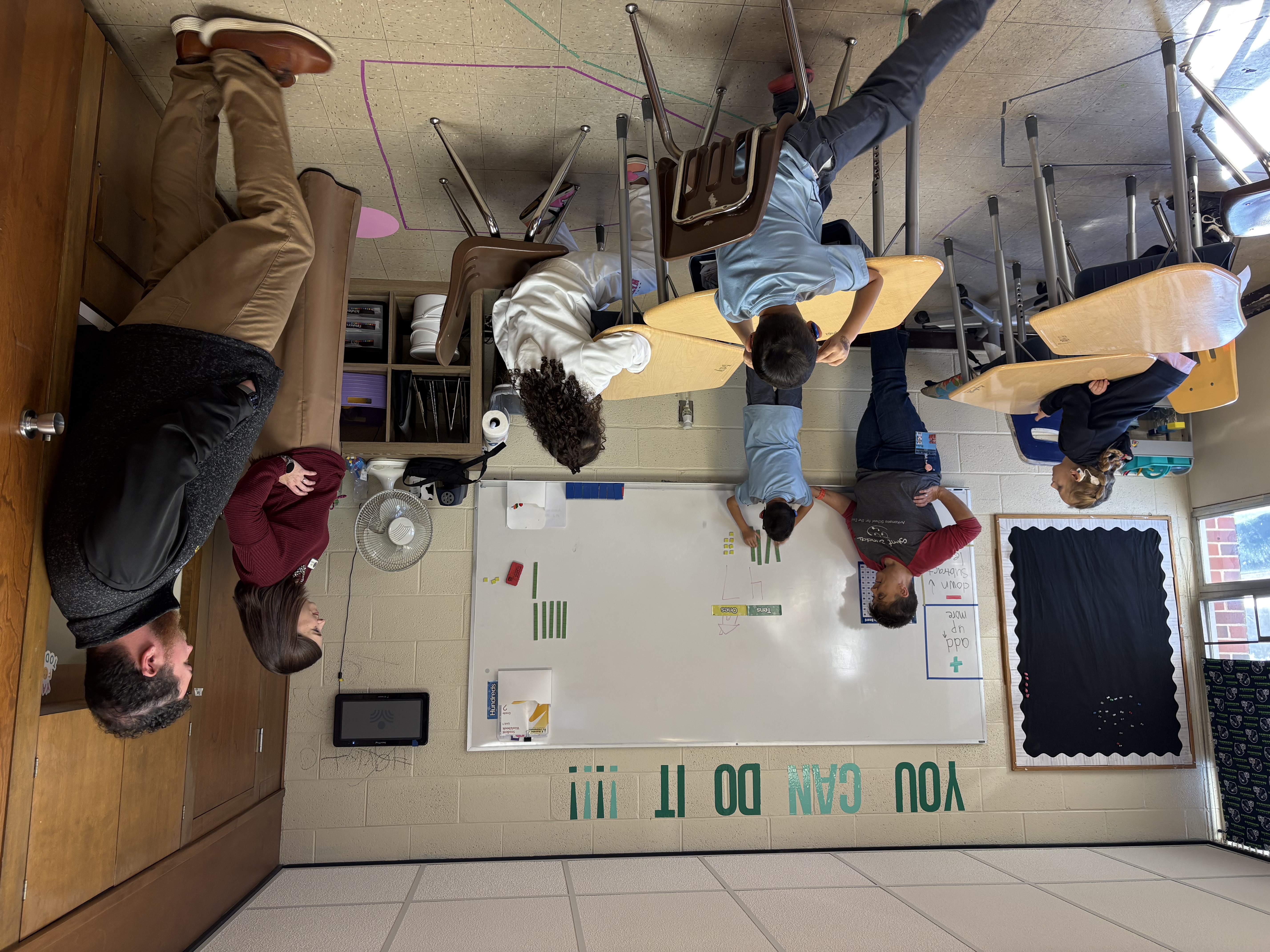 In a classroom setting, a student rearranges number magnets on the white board while other students watch at their desks. Next to him, a teacher watches him, while on the other side of the room two other adults observe the lesson.