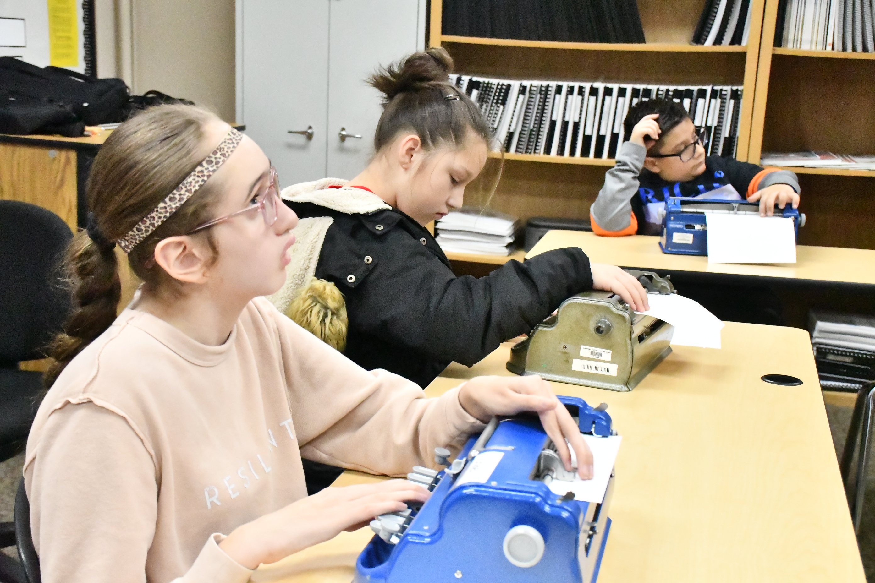 Three students in a classroom setting are seated at desks using Perkins-Braillers.