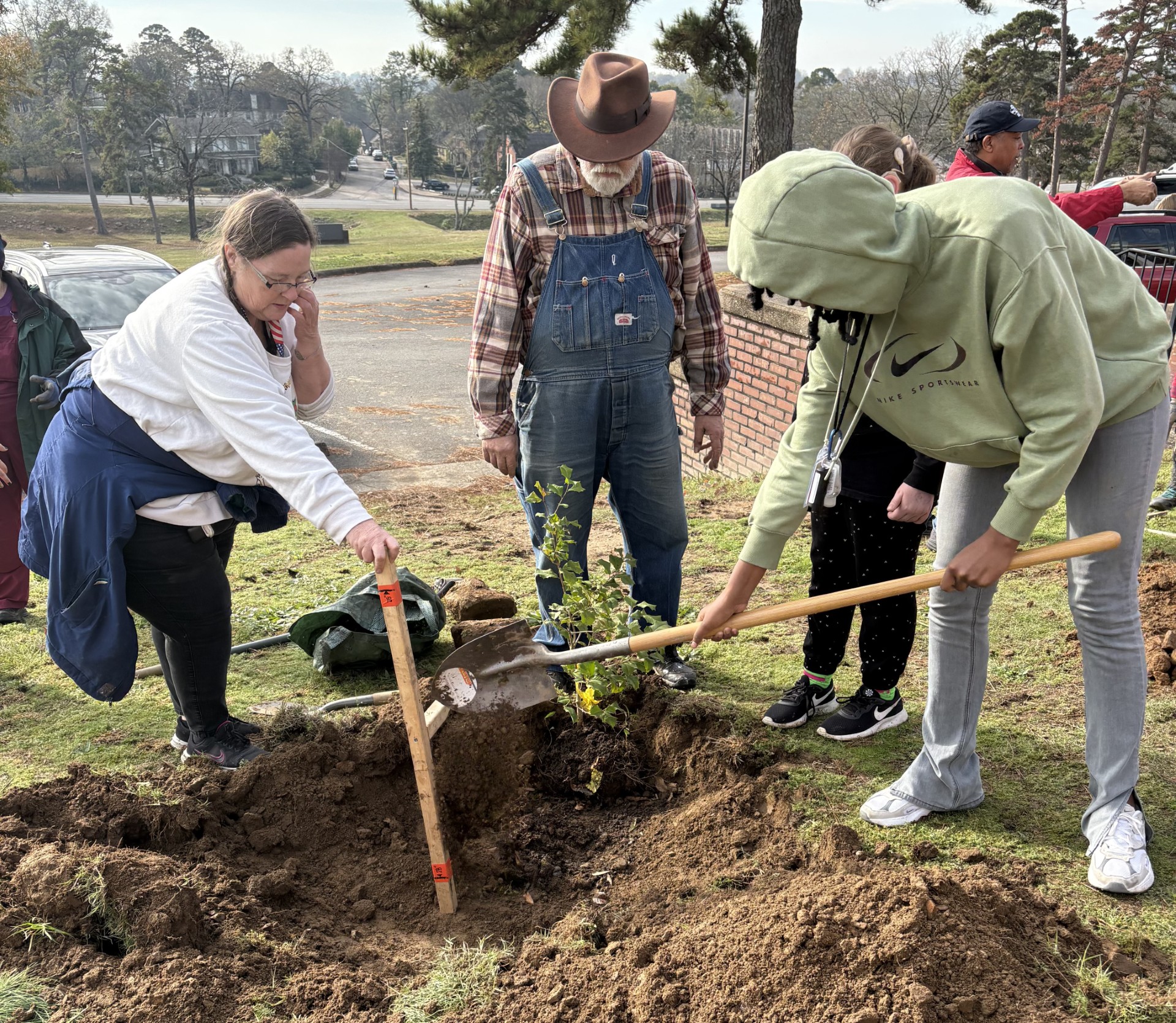 A student shovels dirt over a tree sapling being planted, while volunteers standby to watch and help.