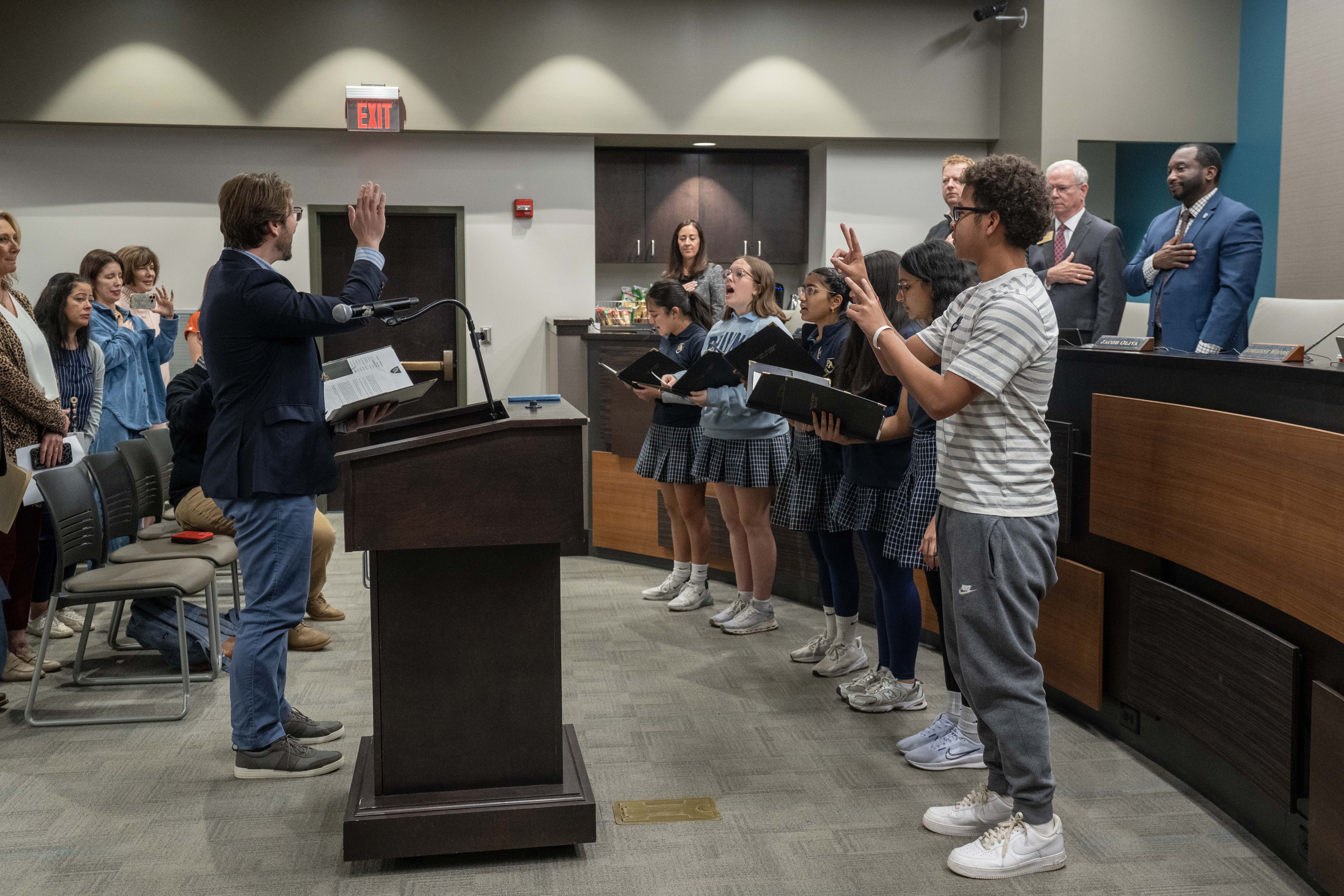On the far right, a student stands next to a group of other students singing in a meeting hall space. The student on the right is signing in ASL, while the students next to him sing while holding open folders.