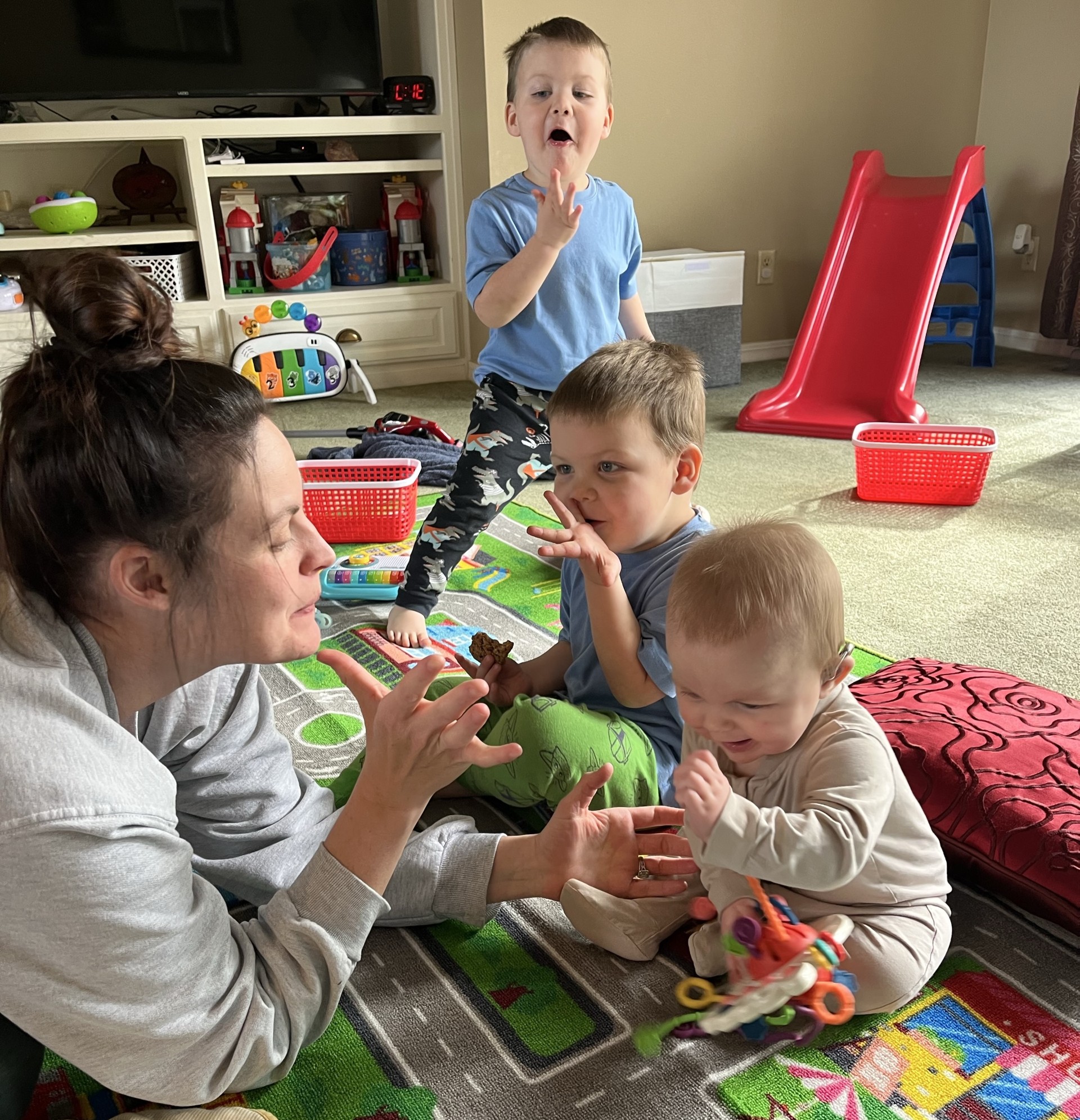 A woman plays on the ground with two toddlers and a baby. The four are practicing signing in ASL together.