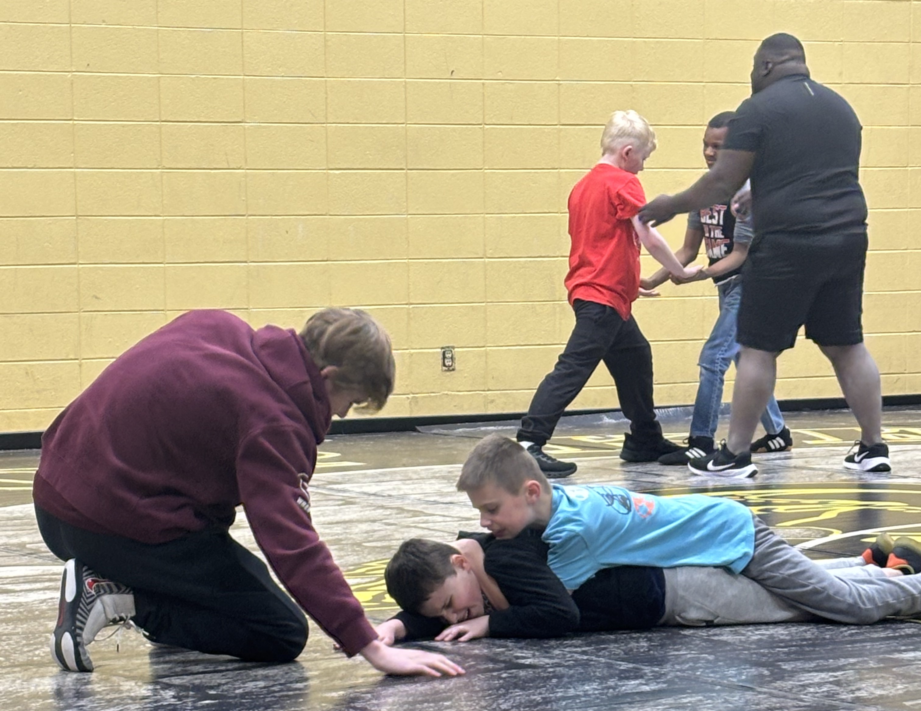 Two students wrestle on the ground while an adult next to them instructs their moves. In the back ground, two more students are beginning to wrestle with another adult helping coach them.